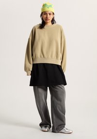 Felpa beige oversized con orlo a costine, indossata sopra una maglietta nera, abbinata a jeans grigi larghi e scarpe argentate lucide. Cappello verde neon con design.