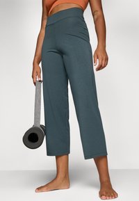 Teal vide-leggede yogabukser laget av myk stoff, med høy midje og en jevn tekstur. Holdt med en grå yogamatte i den andre hånden.