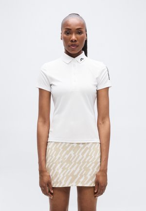 Hvit polo skjorte med krage og knappestolpe, kombinert med et lyst beige skjørt med et abstrakt hvitt mønster.