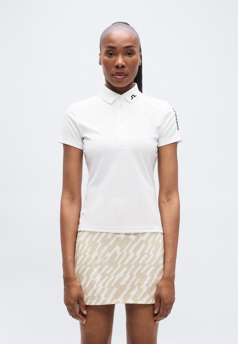 Λευκό πόλο μπλουζάκι με γιακά και λωρίδα με κουμπιά, συνδυασμένο με φούστα ανοιχτού μπεζ χρώματος με αφηρημένο λευκό μοτίβο.