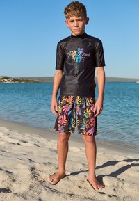Juodas apsauginis marškinėlis su spalvotu užrašu „Palm Beach“ ir juodi maudymosi šortai su ryškiu tropinių lapų raštu, stovintys ant smėlio prie vandens.