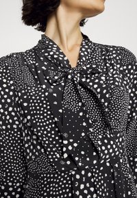 Svart tygklänning med vitt prickmönster, med knytband vid halsen och veckad design, långa ärmar och en mjuk textur.