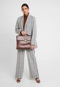 Maletín de cuero marrón con un cierre de metal, forma rectangular, textura suave, sostenido por una persona que lleva un traje de cuadros gris y un suéter de cuello alto marrón.