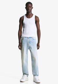 Man staat voorover gericht, draagt een witte tanktop, lichtblauwe jeans, witte sneakers en een dunne ketting, tegen een effen witte achtergrond.