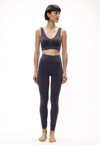Sivá športová podprsenka s nariaseným detailom vpredu, skombinovaná s vysokým pásom sivých legín. Obe vyrobené z hladkej, pružnej látky. Modelka bosá.
