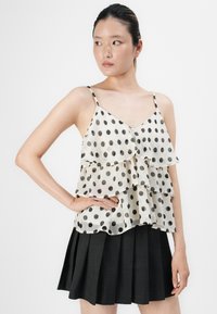 Femme portant un haut sans manches blanc à pois noirs et à volants, associé à une jupe plissée noire, sur un fond uni.