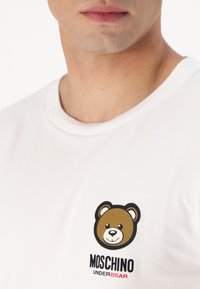 Valge puuvillane t-särk, mis omab ümmargust kaelust, millel on prinditud pruun karu graafika ning musta ja punasega "MOSCHINO UNDERBEAR" tekst.