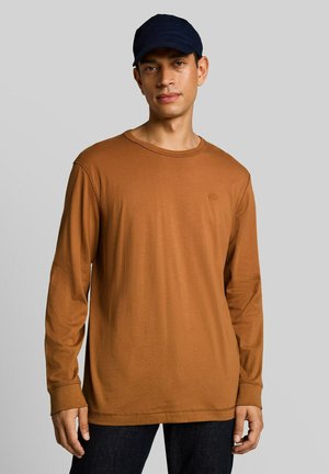 Man met een bruine blouse met lange mouwen en een zwarte pet, staand tegen een effen lichtgrijze achtergrond.