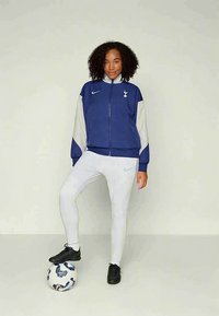 Donna in abbigliamento sportivo blu e bianco si siede con un piede su una palla da calcio contro uno sfondo chiaro e uniforme.