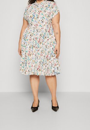 Frau trägt ein knielanges, kurzärmeliges Kleid mit einem mehrfarbigen geometrischen Muster, kombiniert mit schwarzen spitzen Pumps.