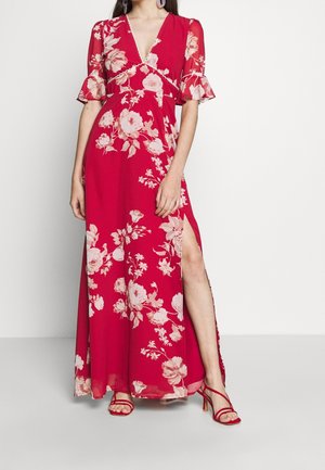 Vrouw draagt een rode maxi jurk met bloemenprint, korte zwierige mouwen en een hoge split, gecombineerd met rode sandalen met hak en bandjes.