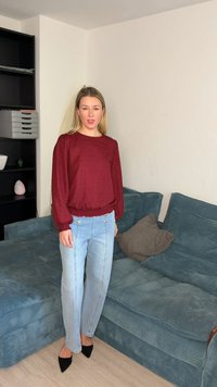 Femme debout à l'intérieur, portant un chemisier bordeaux, un jean bleu clair et des chaussures noires pointues à côté d'un canapé en velours côtelé bleu avec des coussins marron.
