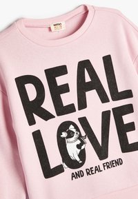 Ljusrosa sweatshirt med fet svart text "REAL LOVE" och en illustration av en hund som håller ett ben. Ribbad rund hals utbildning och ärmar.