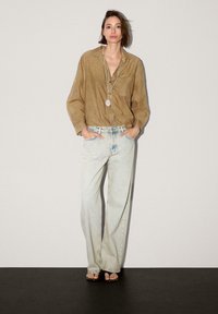 Ljusbrun linnen skjorta med knäppning, lösa blå denimjeans och svarta sandaler. Lång silverhalsband med ett hänge. Enkel bakgrund.
