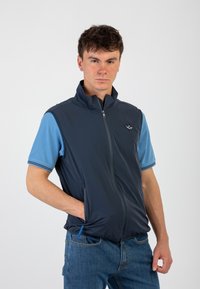 Gilet senza maniche blu navy con zip frontale, tasche e colletto alto, abbinato a una camicia a maniche corte leggera blu chiaro con dettagli a righe.