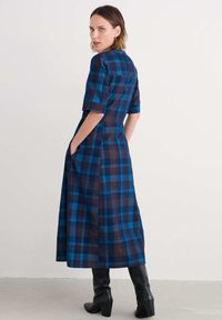 Nő kék és sötét kockás midi ruhában, könyökhosszig érő ujjakkal, keze a zsebében, fekete magassarkú csizmát viselve, egy sima falnak támaszkodva.