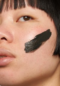 Nærbilde av en kvinnes ansikt med kort svart hår og en tykk stripe med svart krem påført på høyre kinn.