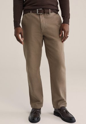 Man draagt een bruine trui, beige broek met riem, donkerbruine loafers met kwastjes en een ring aan zijn rechterhand, staand tegen een lichte achtergrond.