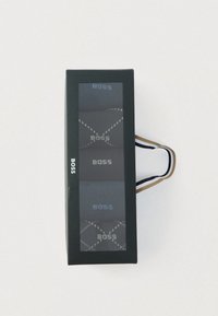 Ensemble emballé de trois paires de chaussettes dans différentes nuances de gris, avec le mot "BOSS" et un motif géométrique. La boîte comporte une fenêtre transparente.