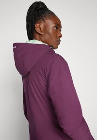Veste imperméable violette avec capuche, dotée d'une doublure vert clair. Tissu texturé, poignets élastiques et cordon de serrage ajustable au niveau du cou.