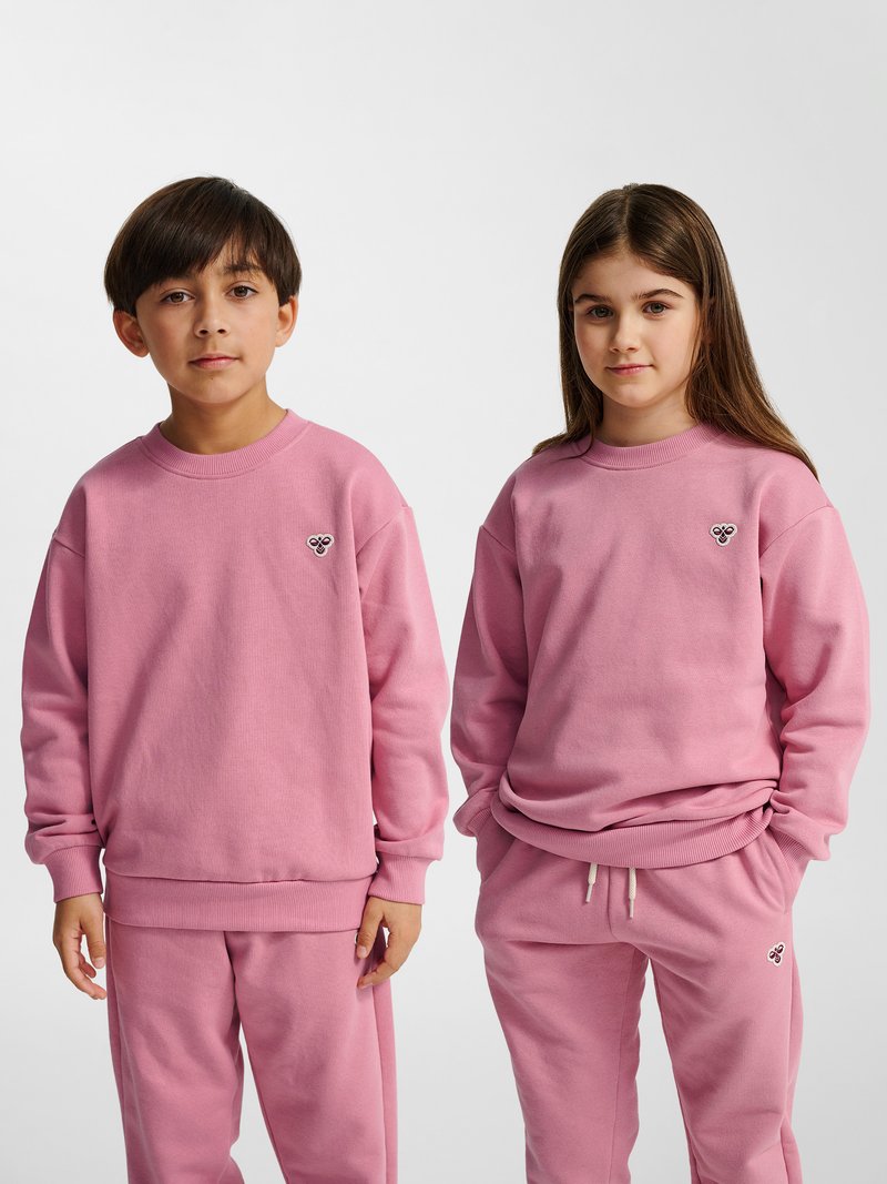 Zwei Kinder stehen nebeneinander und tragen passende pinke Sweatshirts und Hosen mit kleinen herzförmigen Logos auf der linken Brust und am Oberschenkel.
