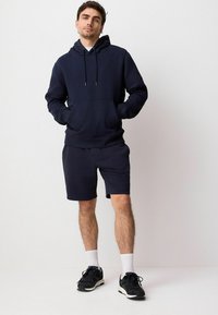 Sudadera de algodón azul marino con un bolsillo frontal, capucha con cordones, combinada con pantalones cortos a juego en azul marino. Zapatillas negras con calcetas blancas completan el conjunto.