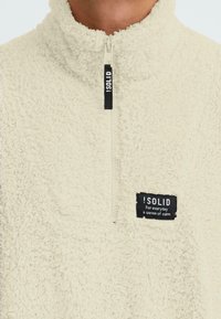 Fleece-Half-Zip-Pullover in Off-White, mit strukturiertem Oberflächen, hohem Kragen und einem schwarzen Etikett mit weißem Text in der Nähe des Reißverschlusses.