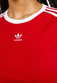 Červené bavlněné tričko s kulatým výstřihem, b�ílými akcenty a třemi bílými pruhy na ramenou. Obsahuje malé bílé logo Adidas.