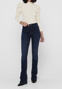 Cream knitted top with puff sleeves, paired with dark blue flared jeans, both featuring a fitted design. Black pointed shoes complete the look.