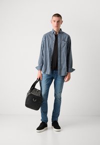 Camisa de cuadros azul y blanca sobre una camiseta negra, combinada con jeans azul claro y zapatillas negras. Sosteniendo una bolsa negra.