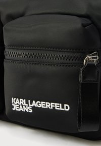 Μαύρη τσάντα από νάιλον με τσέπη με φερμουάρ, με υφή επιφάνειας και λευκό ανάγλυφο λογότυπο "KARL LAGERFELD JEANS" στο μπροστινό μέρος.