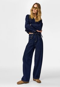 Chaqueta de mezclilla azul oscuro con mangas anchas, combinada con pantalones de pierna ancha a juego. Ambos cuentan con costuras amarillas en contraste. Calzado marrón completa el conjunto.