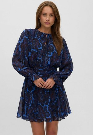 Mujer que lleva un vestido de manga larga con estampado paisley azul y negro, con un cuello alto y puños con volantes, de pie con las manos entrelazadas.