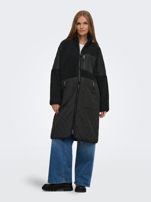 Donna in piedi che indossa un lungo cappotto nero trapuntato e in pile, jeans blu a gamba larga e stivali neri robusti su sfondo bianco.