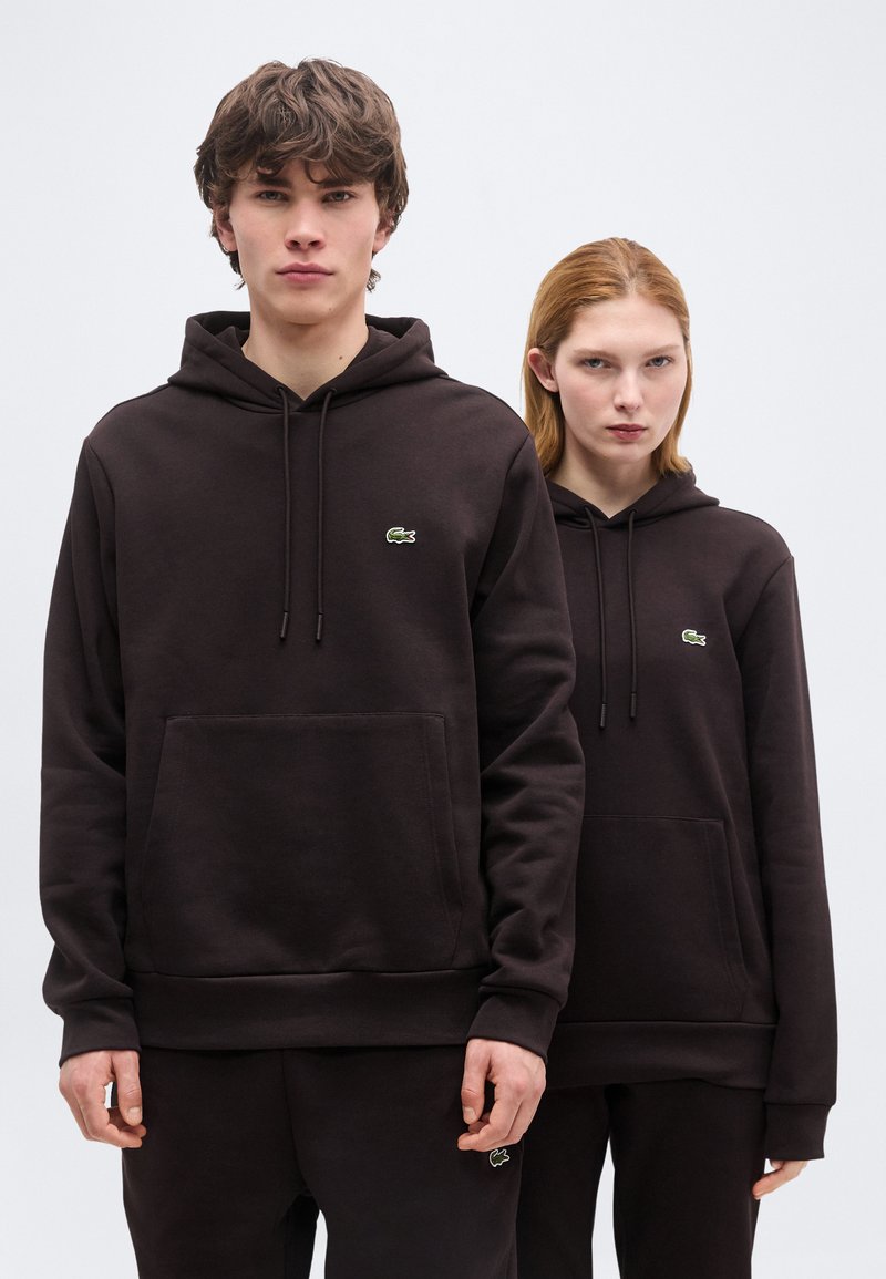 Νεαρός άνδρας και γυναίκα που φορούν μαύρα φούτερ με κουκούλα και αντίστοιχο παντελόνι, στέκονται μπροστά σε απλό λευκό φόντο.