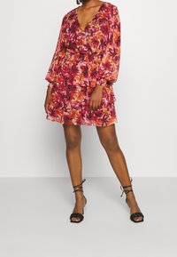 Vestido mini floral con escote en V, mangas largas y cintura fruncida, con tonos en rosa, rojo y naranja. Sandalias de tiras negras.
