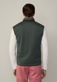 Grüne gesteppte Weste mit strukturierter Oberfläche, braunem Kragen und seitlichen Knopf-Akzenten. Getragen über einem weißen Langarmshirt und rosa Hosen.