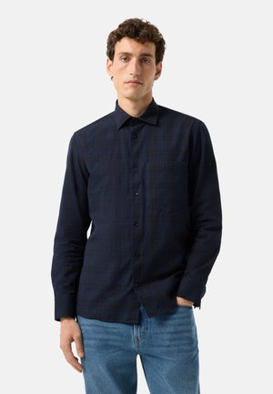 Mann mit kurzen lockigen Haaren, der ein dunkelblaues Hemd mit dezentem Karomuster und blaue Jeans trägt, steht vor einem schlichten hellen Hintergrund.