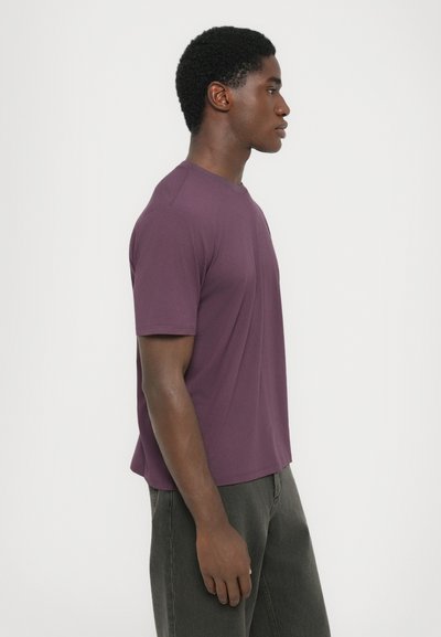 Young man in purple short-sleeve t-shirt and dark pants standing in profile against a plain light background.