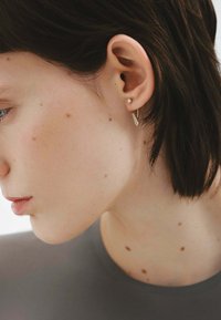 Aro de oro con superficie texturizada; pequeño arete de botón redondo. El cabello corto del modelo enmarca parcialmente los aretes y la oreja.