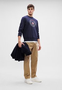 Granatowy sweter z okrągłym nadrukiem z przodu, beżowe spodnie z kordurowej tkaniny i białe sneakersy, trzymane z złożonym granatowym swetrem.