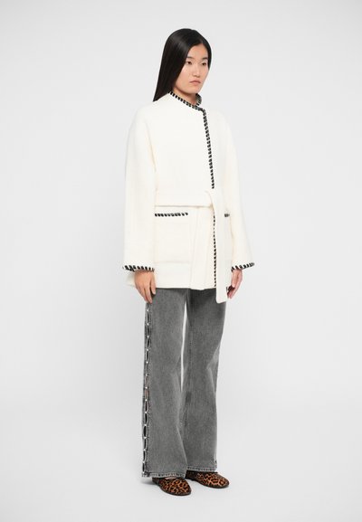 Manteau en fausse fourrure blanche avec des finitions noires, ceinturé à la taille, associé à un jean gris avec des détails sur les côtés et des chaussures à enfiler à imprimé animal.