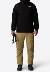 Schwarze wasserdichte Jacke mit Reißverschluss, khakifarbene Cargo-Hosen mit Reißverschlusstaschen und dunkelblaue Wanderschuhe. Person hält schwarze Trekkingstöcke.