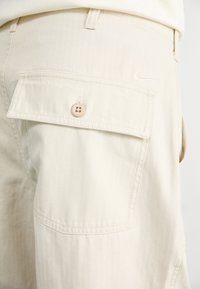 Bermudas bejes em tecido de sarja com textura de espinha de peixe, um bolso com aba e fecho por botão, e um pequeno logótipo bordado na cintura.