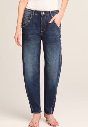 Woman wearing dark blue relaxed-fit jeans with front pockets and beige heeled sandals, standing against plain background.