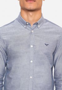 Chemise boutonnée en tissu bleu clair, avec col, manches longues et petit logo brodé sur la poitrine gauche. Coupe classique.