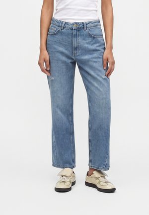 Person iført lyseblå lige jeans med let distress og cremefarvede sneakers med sorte såler, stående mod en ensfarvet baggrund.