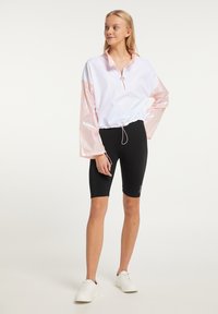 Hvid halv-zip pullover med skinnende pink ærmer, indsnøret talje, parret med sorte cykelshorts og hvide sneakers. Glat stoftekstur.