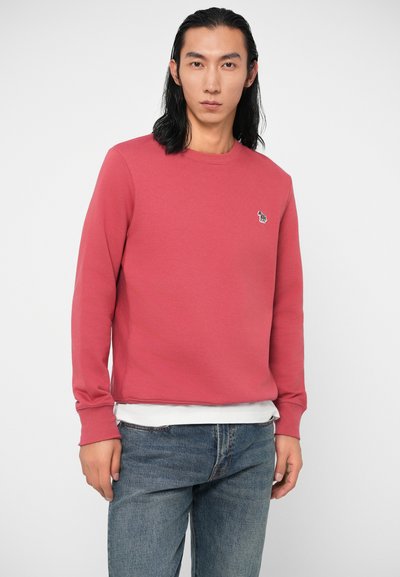 Jaunas vyras su ilgais juodais plaukais, apsivilkęs raudoną džemperį su apvalia iškirpte virš baltos marškinėlių ir mėlynų džinsų, stovi prieš paprastą foną.