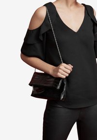 Schwarze Handtasche aus Kunstleder im Krokodil-Design mit Klappenverschluss und silberner Kettenriemen. Kombiniert mit einem schwarzen Oberteil mit kalten Schulterärmeln.